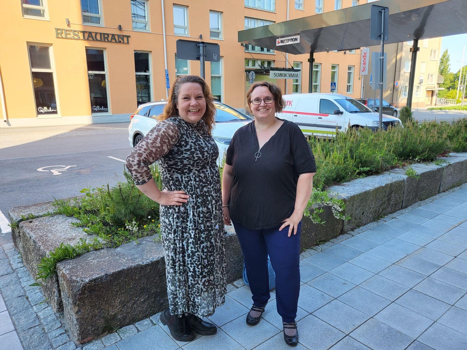 Two people standing on a sidewalk with foliage, with a background of a restaurant and parked cars.